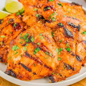 Grilled chicken thighs with a reddish-orange marinade, garnished with chopped herbs, served on a white plate with lime wedges in the background.