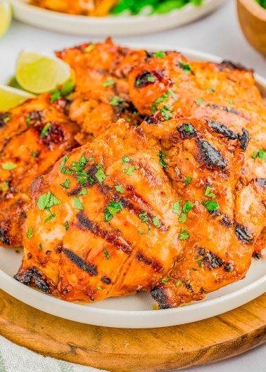 Grilled chicken thighs with a reddish-orange marinade, garnished with chopped herbs, served on a white plate with lime wedges in the background.