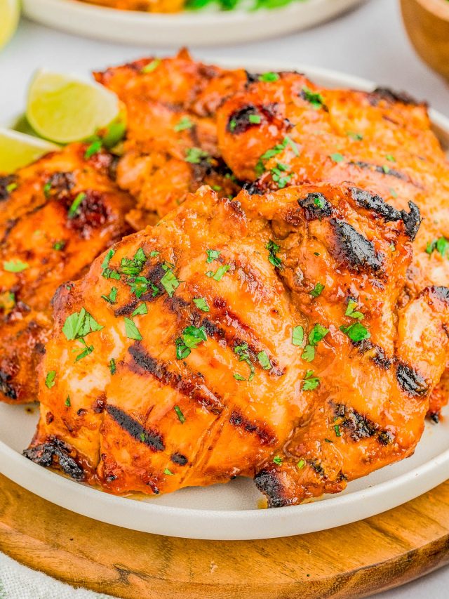 Grilled chicken thighs with a reddish-orange marinade, garnished with chopped herbs, served on a white plate with lime wedges in the background.
