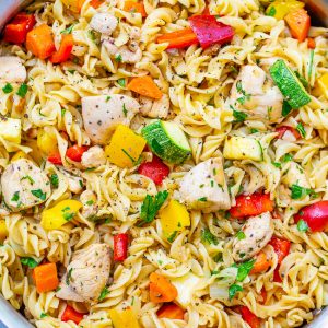 A skillet filled with rotini pasta, diced chicken, and mixed vegetables including zucchini, bell peppers, and carrots, garnished with chopped herbs.