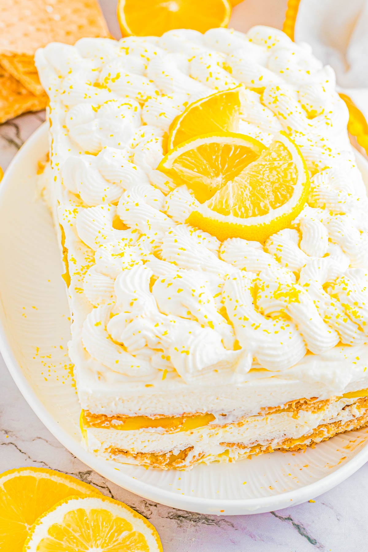 A no-bake lemon icebox cake with layers of whipped cream, lemon slices, and graham crackers, topped with swirls of cream and lemon zest on a white plate.