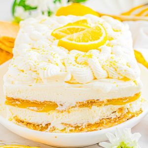 Rectangular lemon dessert with cream layers and lemon slices on top, served on a white plate with lemon slices and white flowers nearby.