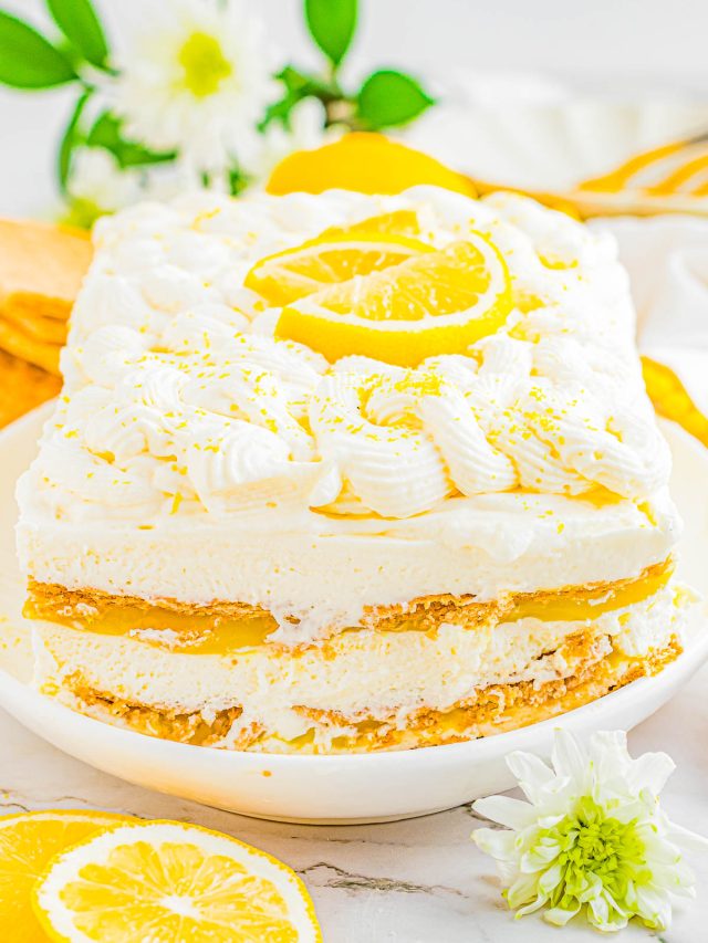 Rectangular lemon dessert with cream layers and lemon slices on top, served on a white plate with lemon slices and white flowers nearby.