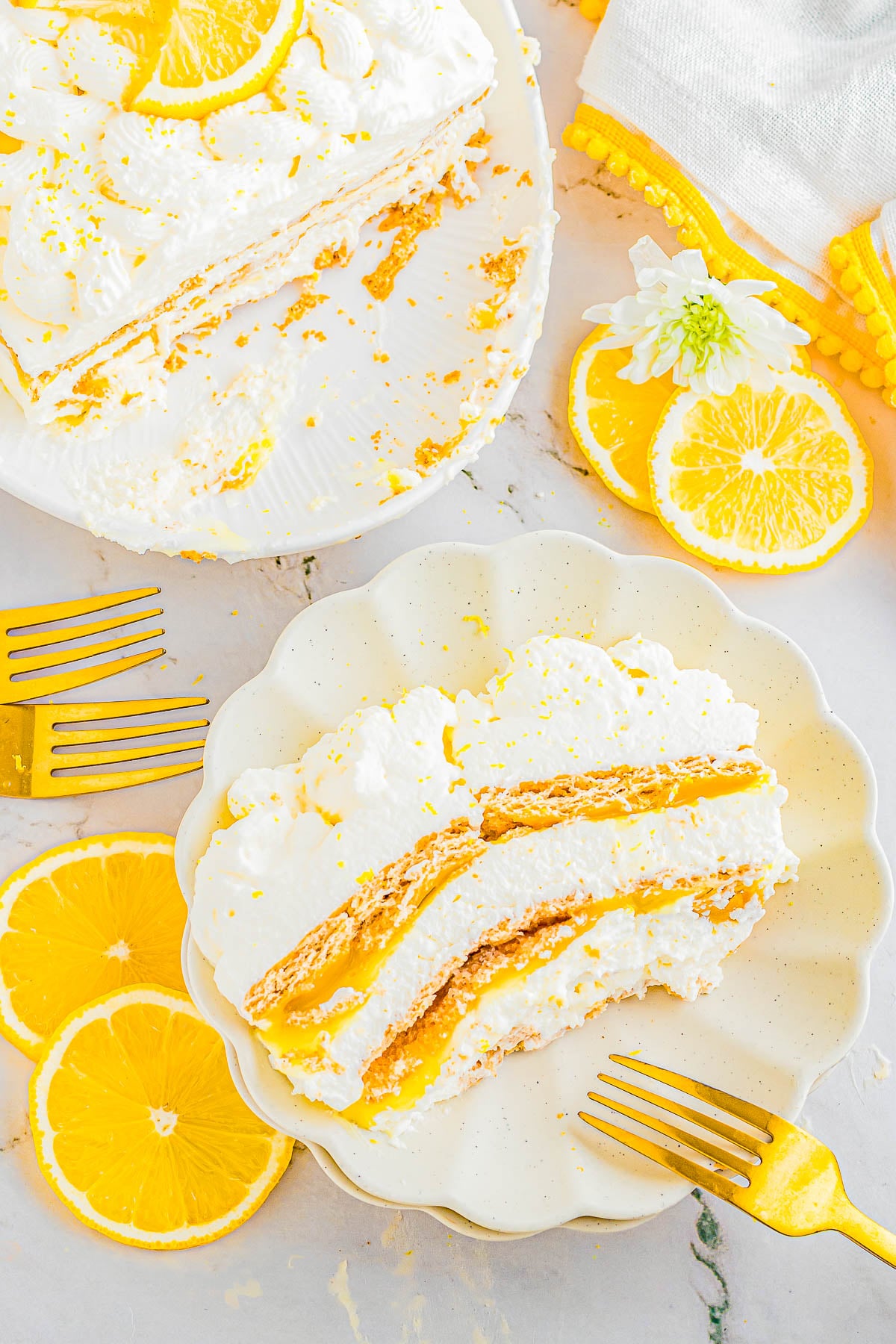 A slice of layered lemon dessert with whipped cream and crust on a plate, surrounded by orange slices and gold forks, with more dessert on a serving dish in the background.