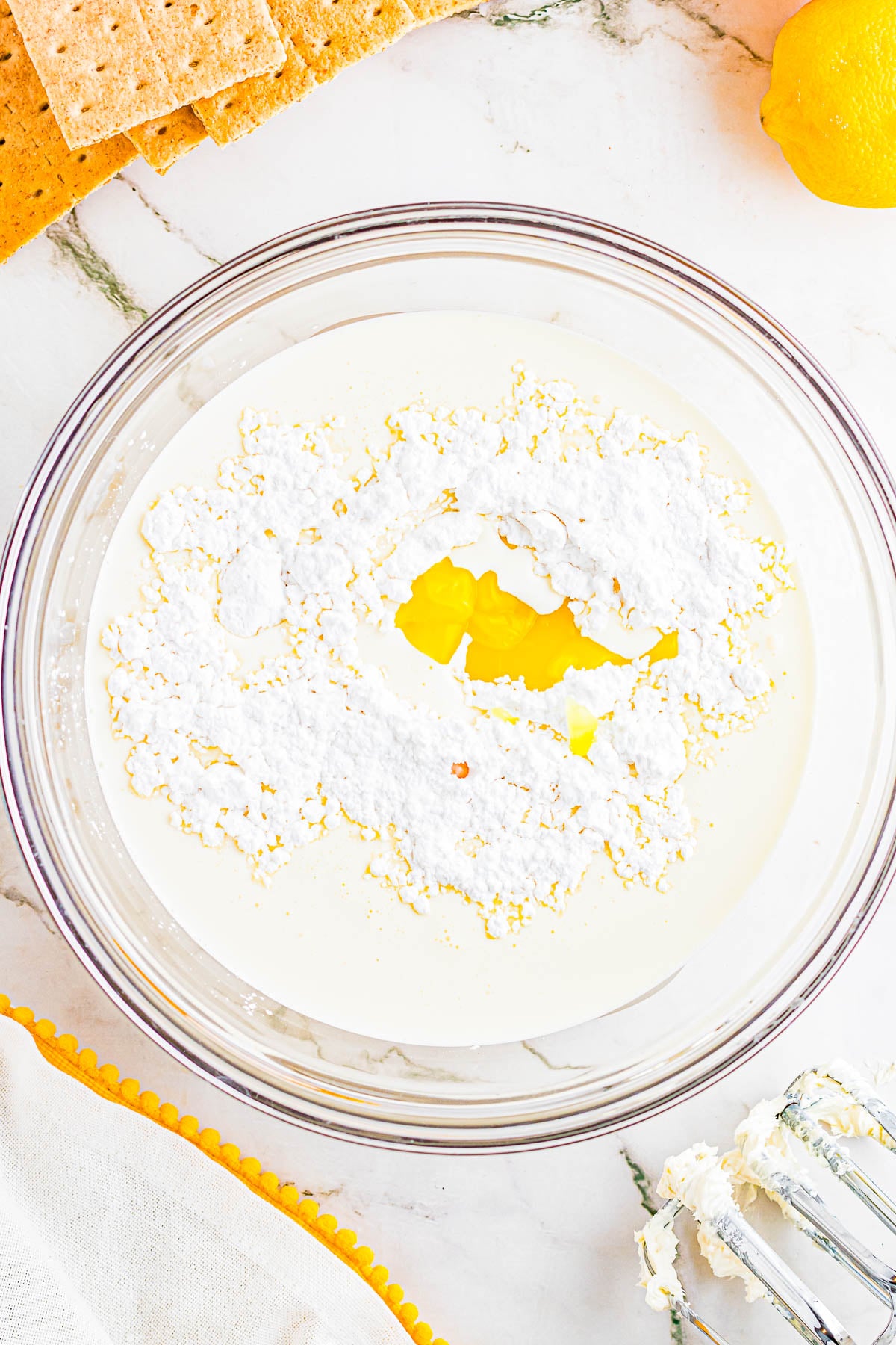 A glass bowl contains cream, powdered sugar, and an egg yolk on a white marble countertop, with graham crackers, a lemon, and a mixer nearby.