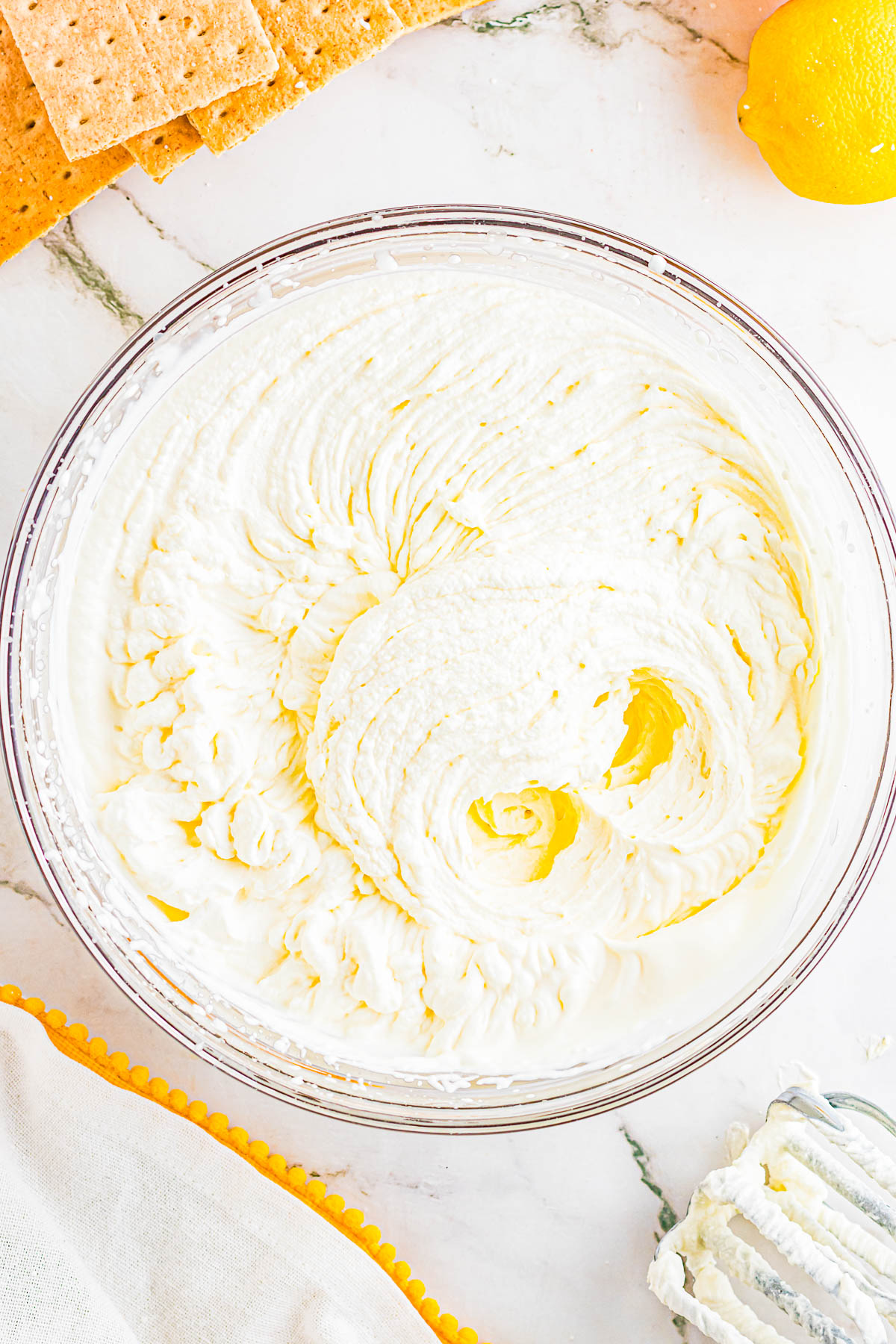 A glass bowl filled with whipped cream sits on a marble surface, surrounded by graham crackers, a lemon, and a cloth napkin.