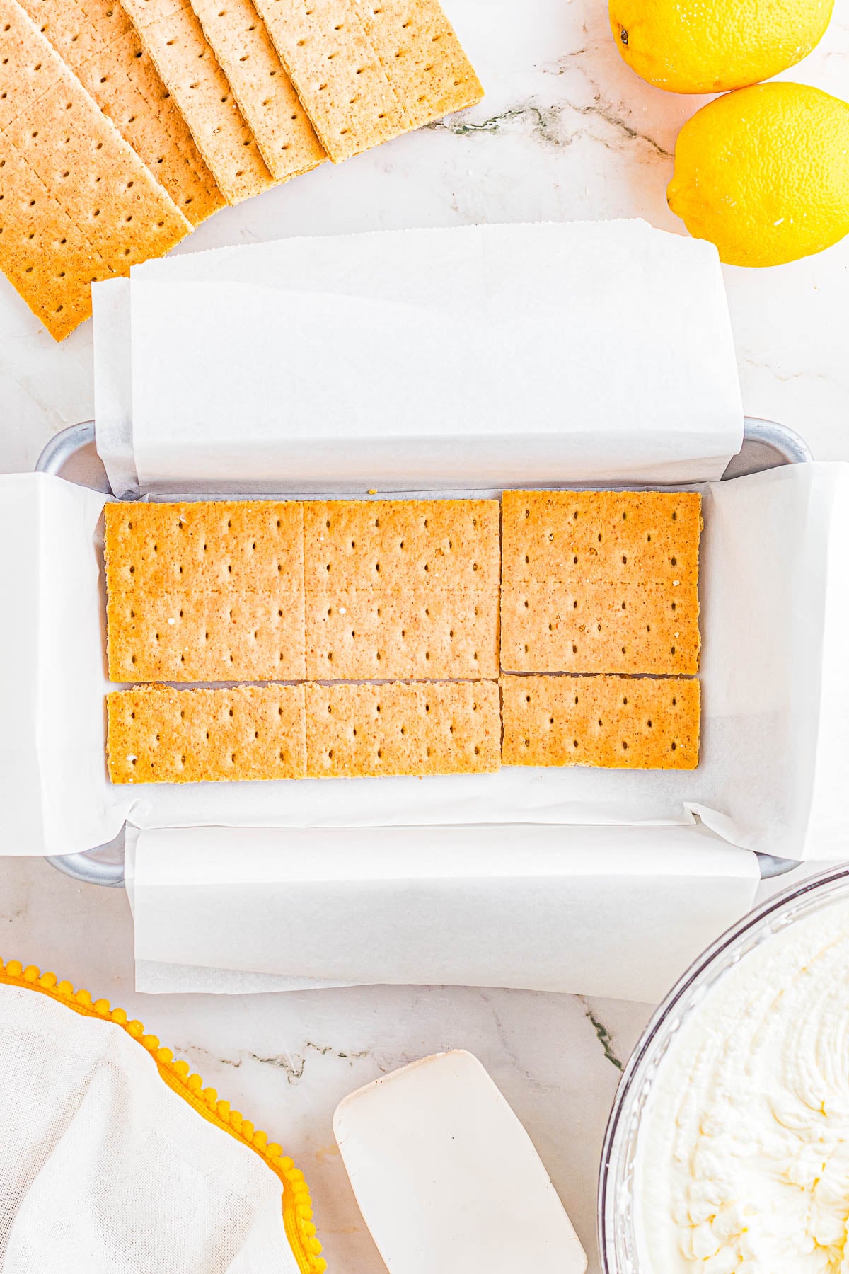 Rectangular pan lined with parchment paper, layered with graham crackers. Additional graham crackers, lemons, a bowl of cream, and a spatula are nearby on a marble surface.