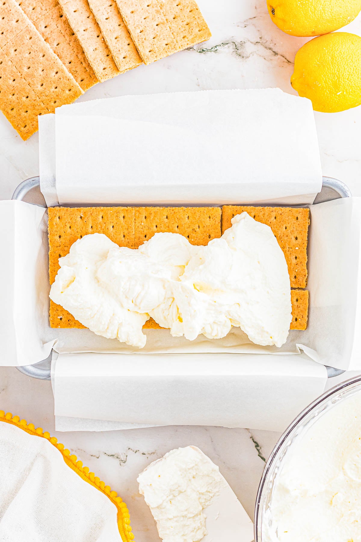 A loaf pan lined with parchment paper contains a layer of graham crackers topped with a layer of whipped cream, with lemons and more graham crackers nearby.