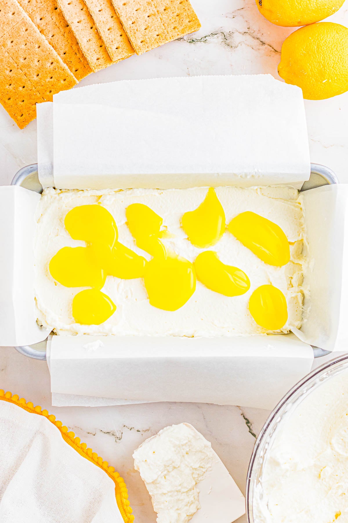 A parchment-lined pan filled with creamy cheesecake mixture, topped with dollops of yellow lemon curd, surrounded by graham crackers and lemons on a marble surface.