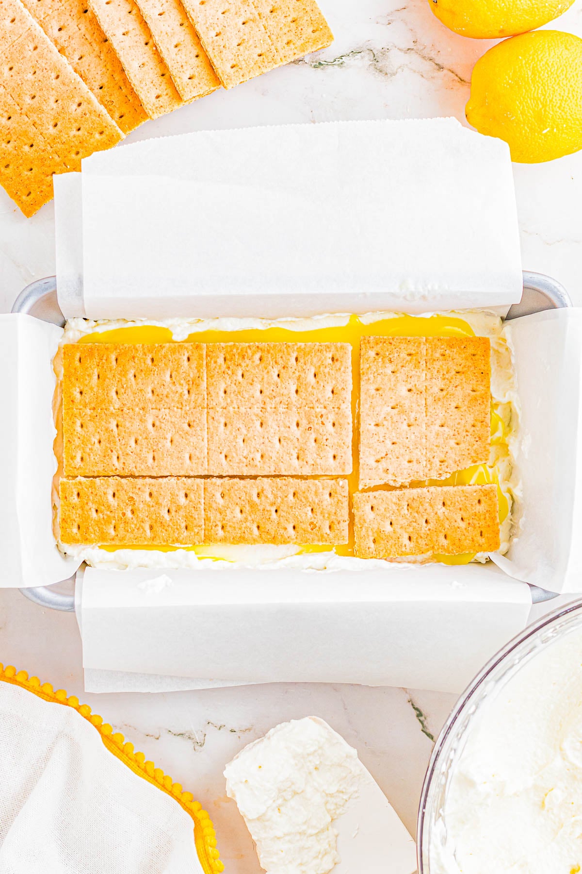 A baking pan lined with parchment paper contains a dessert topped with rectangular graham crackers; lemons and cream are visible nearby.