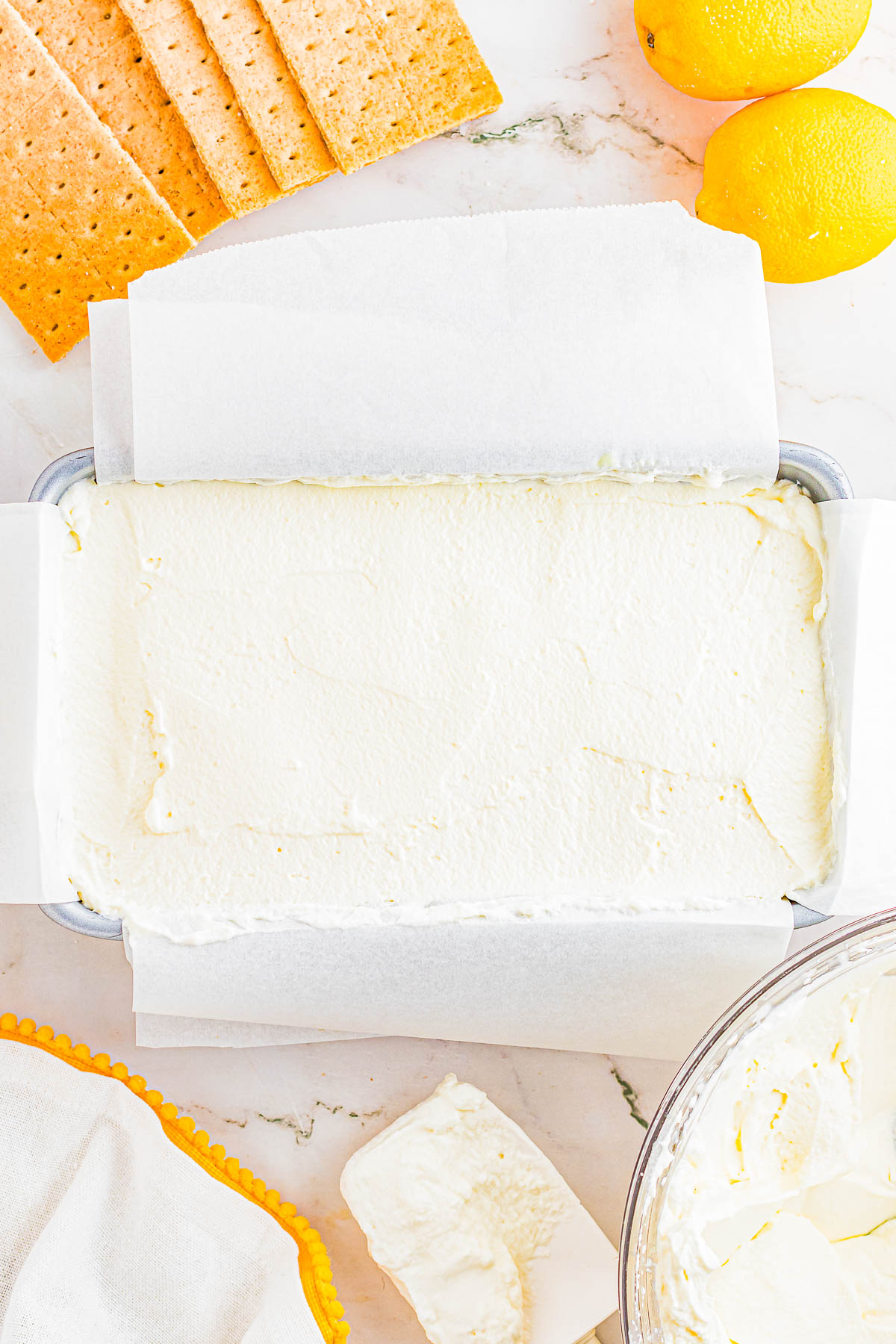 A loaf pan lined with parchment paper is filled with a creamy white mixture. Nearby are graham crackers, lemons, and a block of cream cheese on a marble surface.