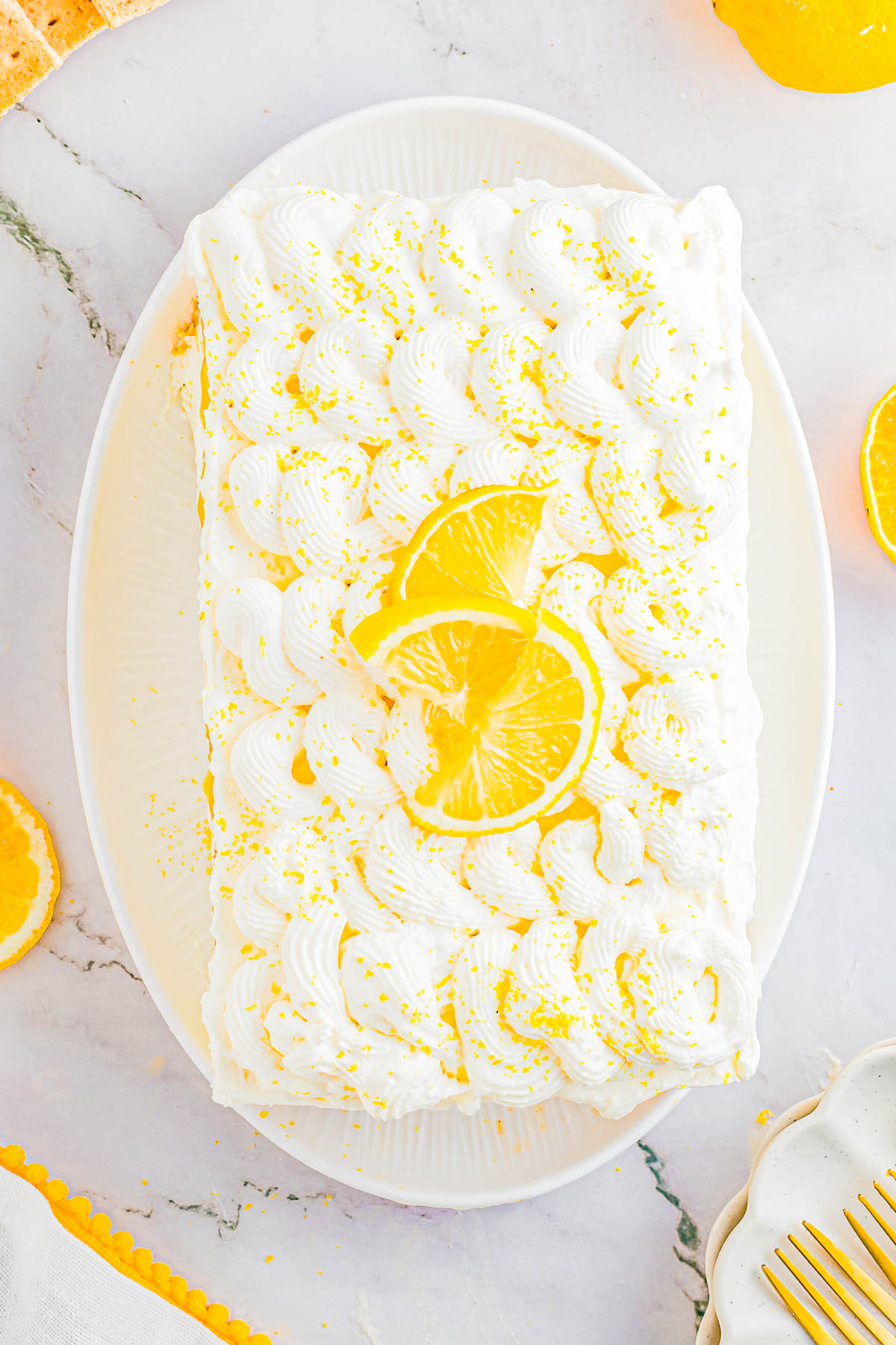 Rectangular lemon cake topped with swirls of white frosting, lemon slices, and yellow sprinkles, displayed on a white oval plate.