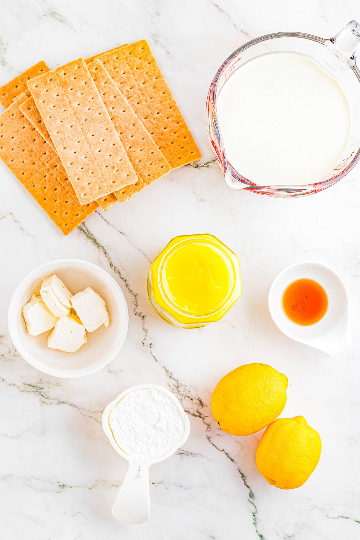 Graham crackers, cream, lemon curd, vanilla extract, lemons, powdered sugar, and cream cheese arranged on a marble surface.