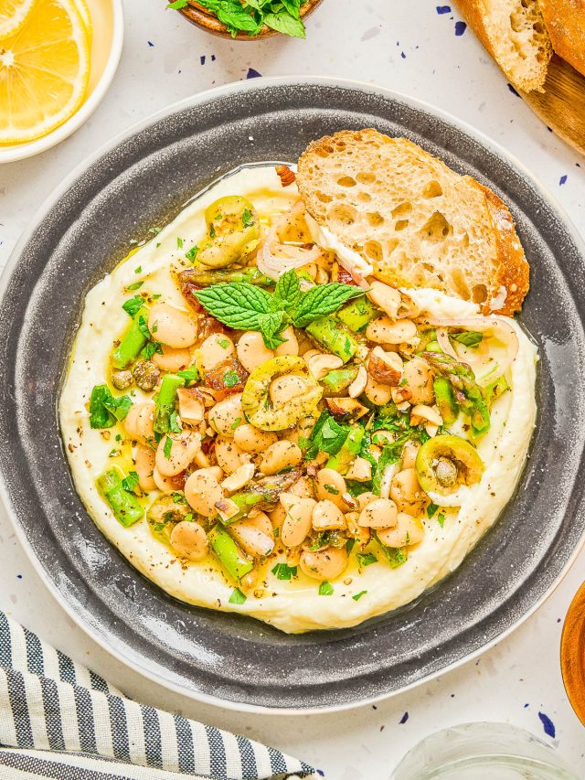 A plate of creamy white hummus topped with chopped green olives, white beans, herbs, and spices, served with a slice of crusty bread.