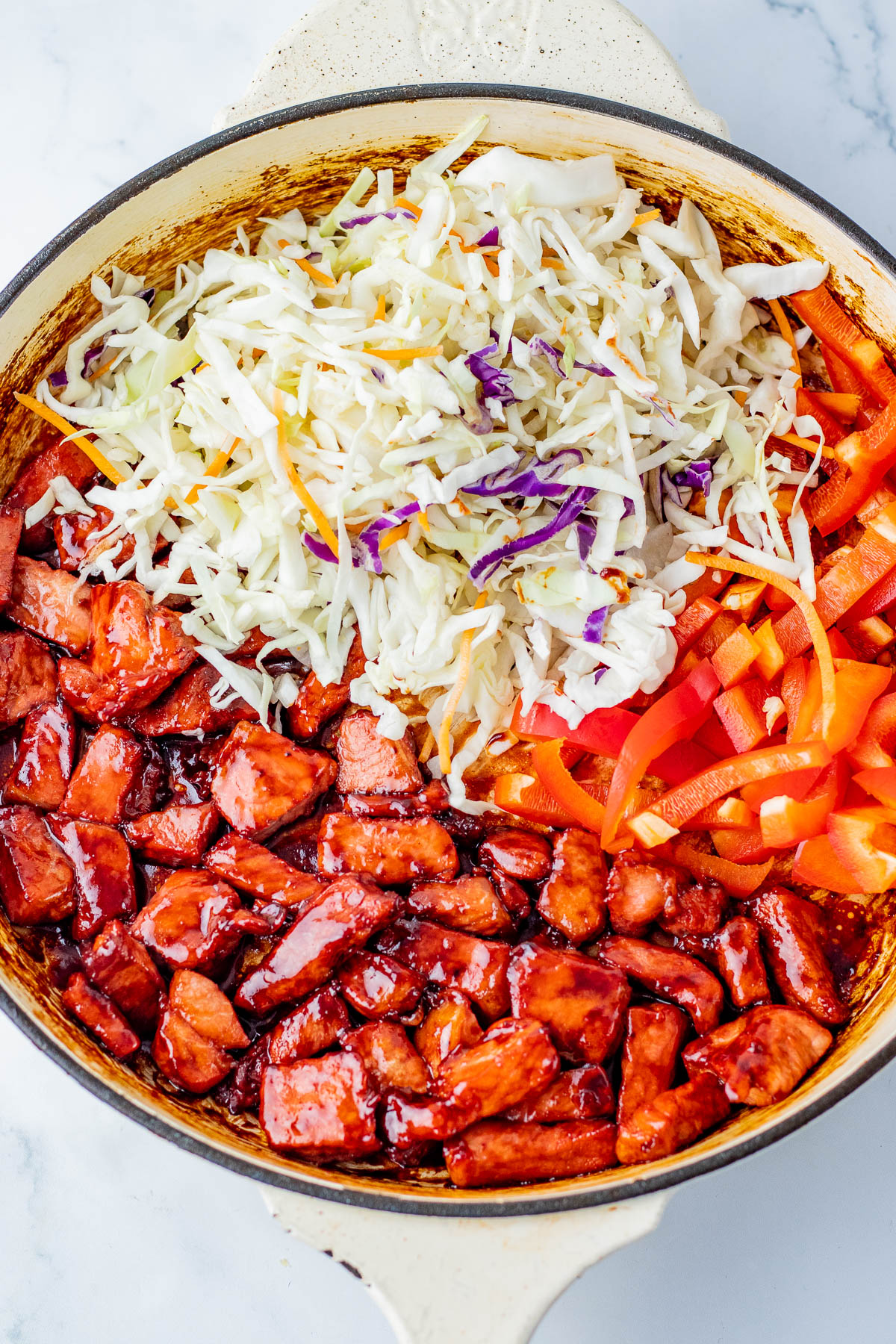A skillet containing cooked chicken in sauce, shredded cabbage, and sliced red bell peppers, with the vegetables uncooked and separated from the chicken.