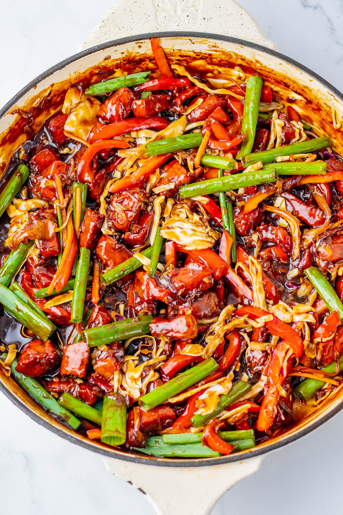 A skillet filled with stir-fried vegetables, including green onions, red bell peppers, carrots, cabbage, and sauce.