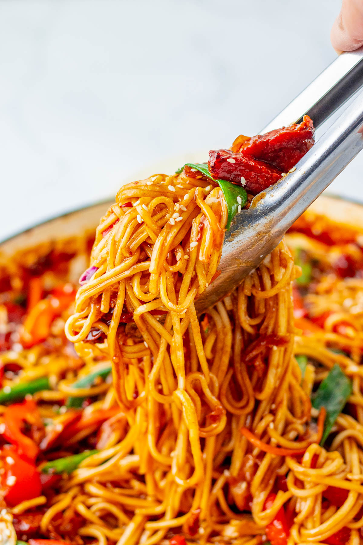 Tongs lifting a serving of stir-fried noodles with vegetables, red chilies, and sesame seeds from a pan.