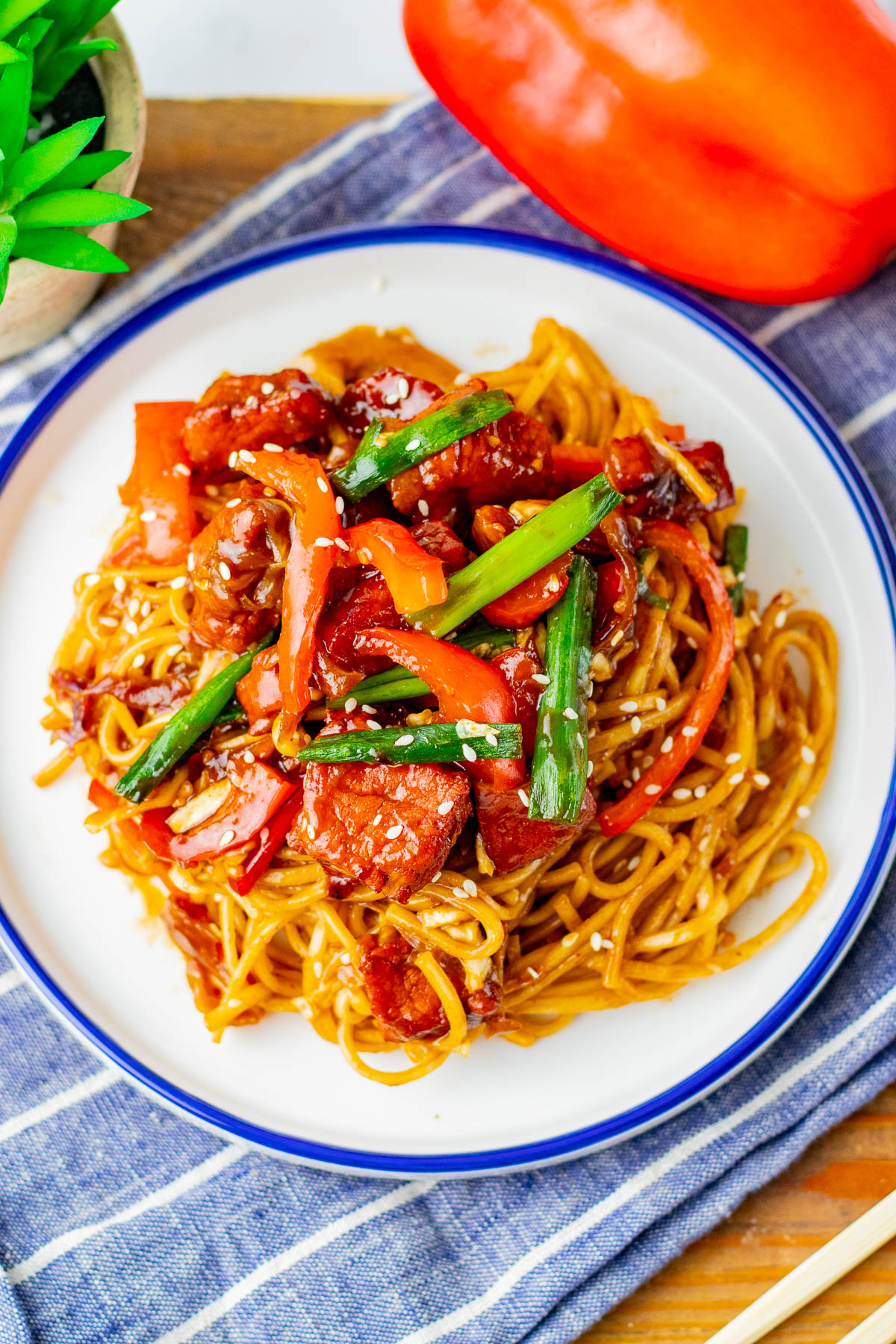 A plate of stir-fried noodles with sliced sausage, red bell peppers, green onions, and sesame seeds, served on a white dish with a blue rim.