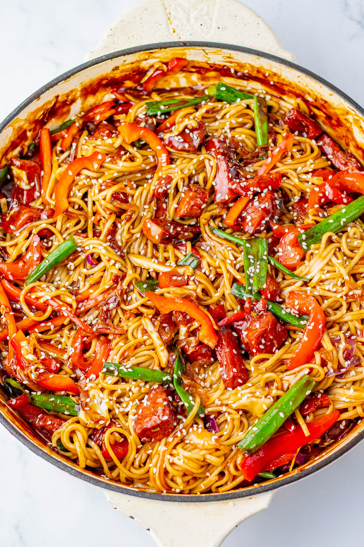 A skillet filled with stir-fried noodles, sliced red bell peppers, green beans, and pieces of meat, topped with sesame seeds.