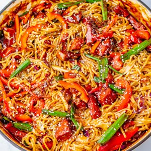 A skillet filled with cooked noodles, sliced red peppers, green beans, sauce, and sesame seeds on top.