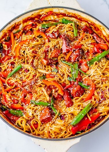 A skillet filled with cooked noodles, sliced red peppers, green beans, sauce, and sesame seeds on top.