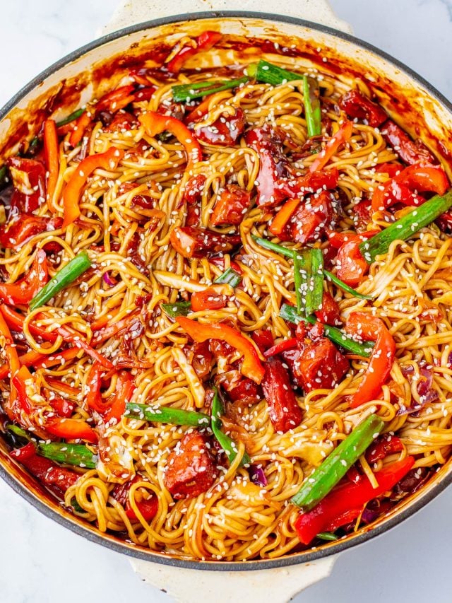 A skillet filled with cooked noodles, sliced red peppers, green beans, sauce, and sesame seeds on top.