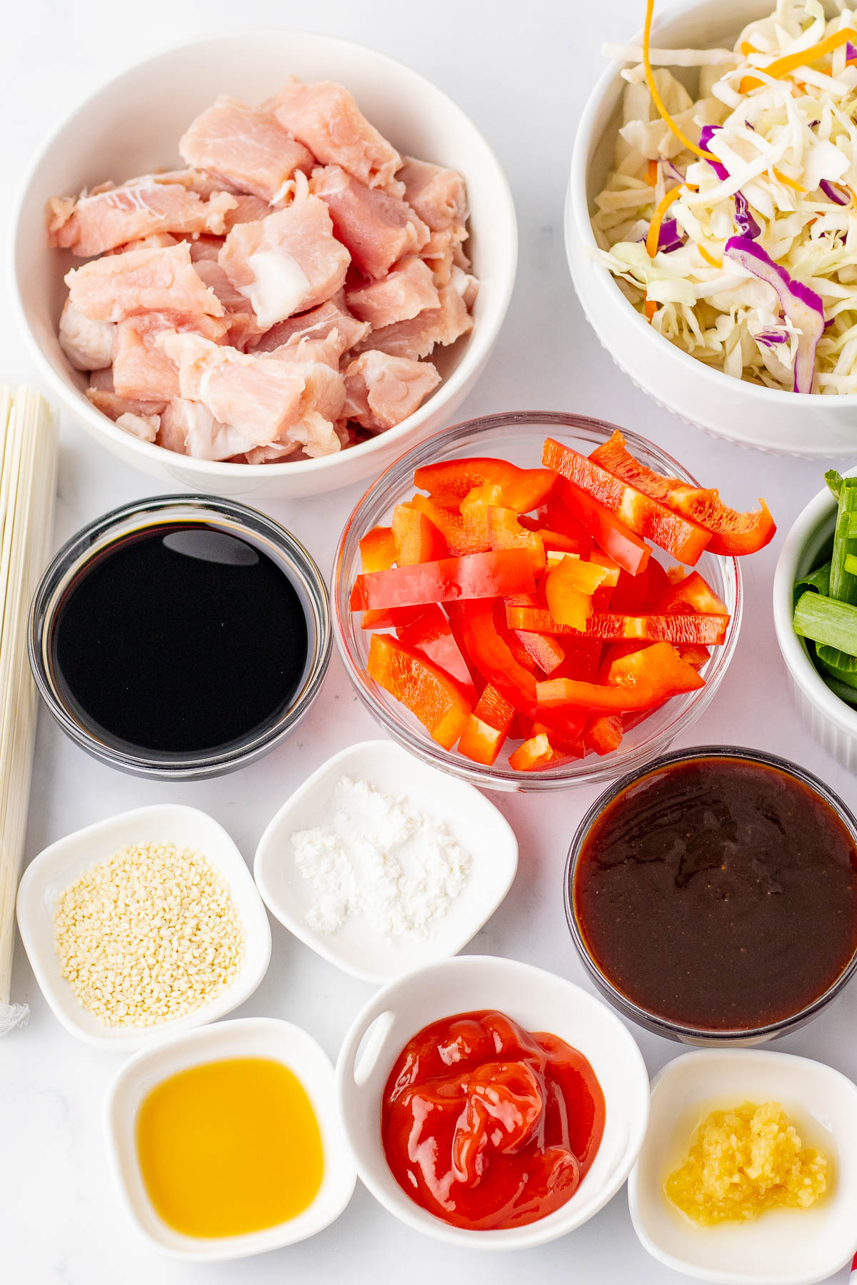 Bowls containing raw chicken, shredded cabbage, sliced red bell pepper, soy sauce, hoisin sauce, sesame seeds, ketchup, minced garlic, cornstarch, oil, and green onions on a white surface.