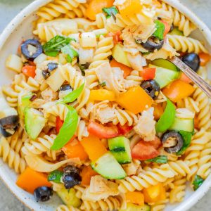 A bowl of rotini pasta salad with cucumbers, tomatoes, yellow bell peppers, black olives, onions, and basil, with a fork and a lemon slice nearby.