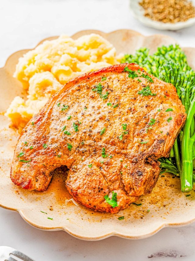 A cooked pork chop garnished with herbs is served on a plate with mashed potatoes and steamed broccolini.