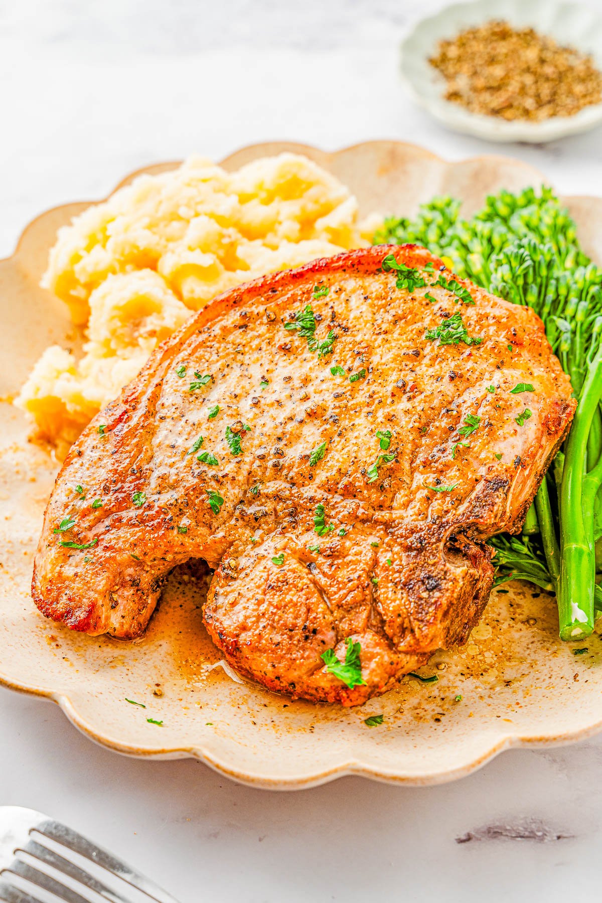 A cooked pork chop garnished with herbs is served on a plate with mashed potatoes and steamed broccolini.