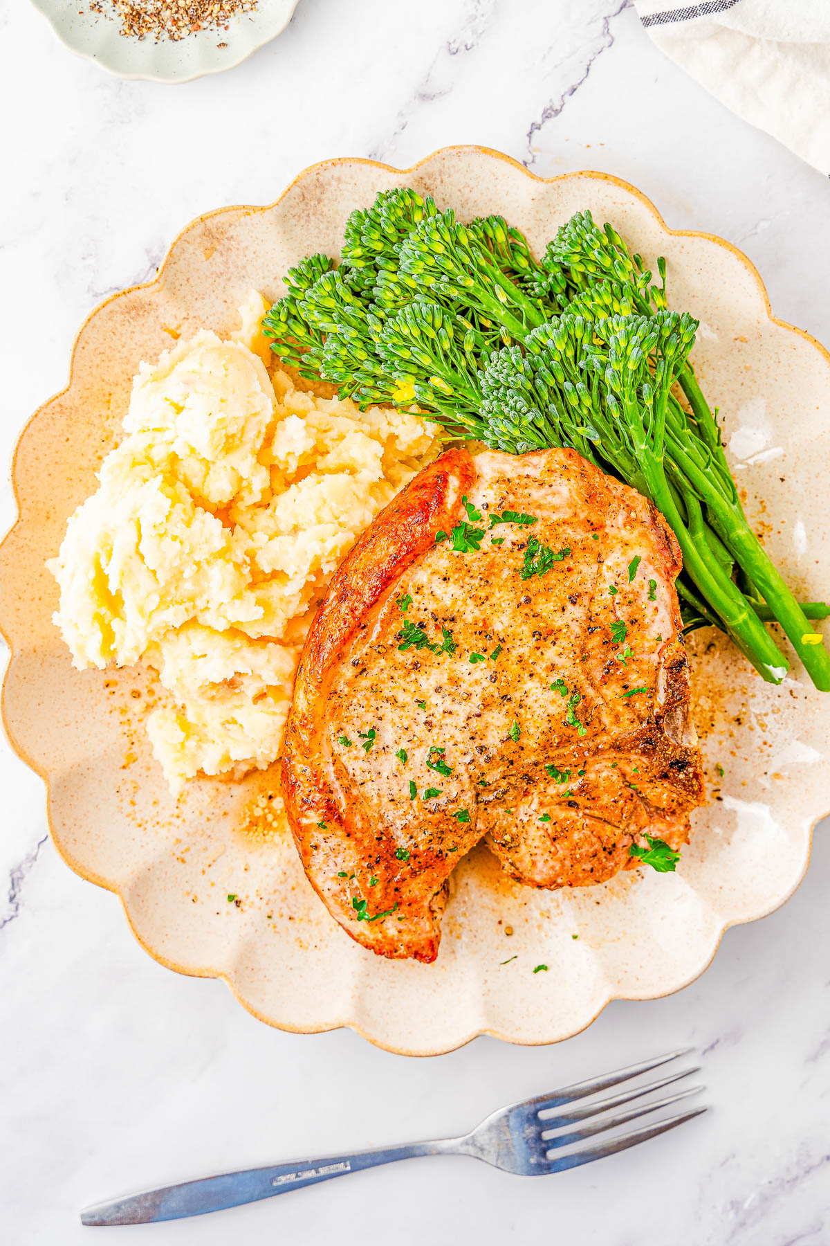 A plate with a seasoned pork chop, mashed potatoes, and steamed broccolini. A fork is placed beside the plate on a white marble surface.