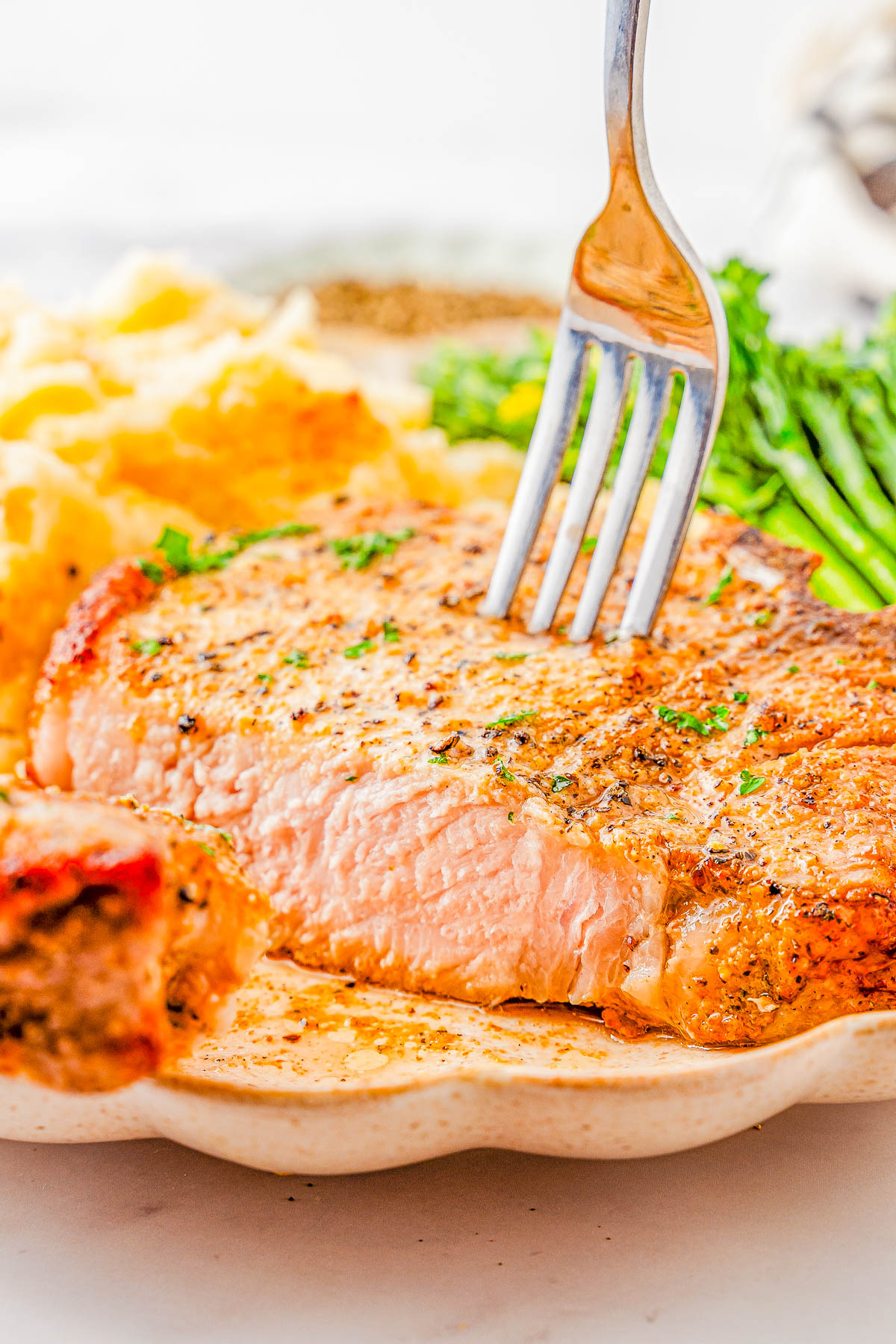 A fork is inserted into a cooked pork chop on a plate with mashed potatoes and green vegetables in the background.