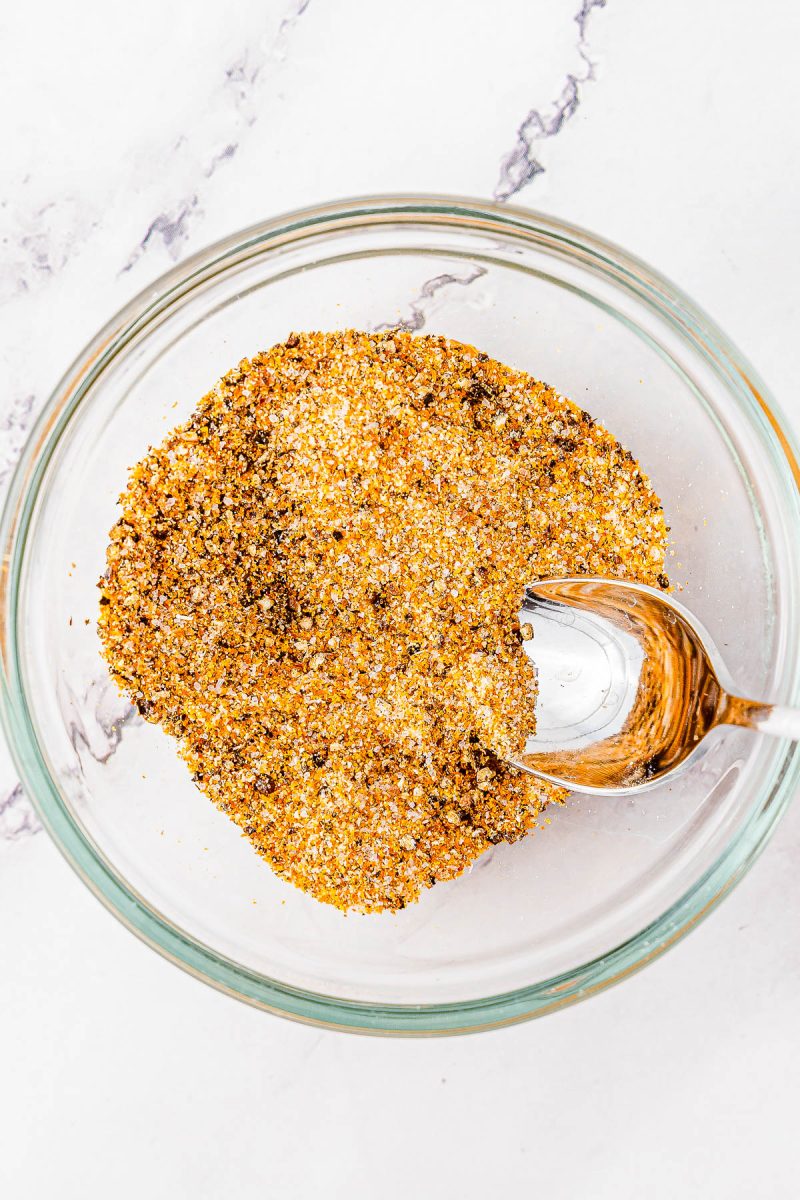 A clear glass bowl filled with a mixed seasoning blend, with a spoon resting inside, on a white marble surface.