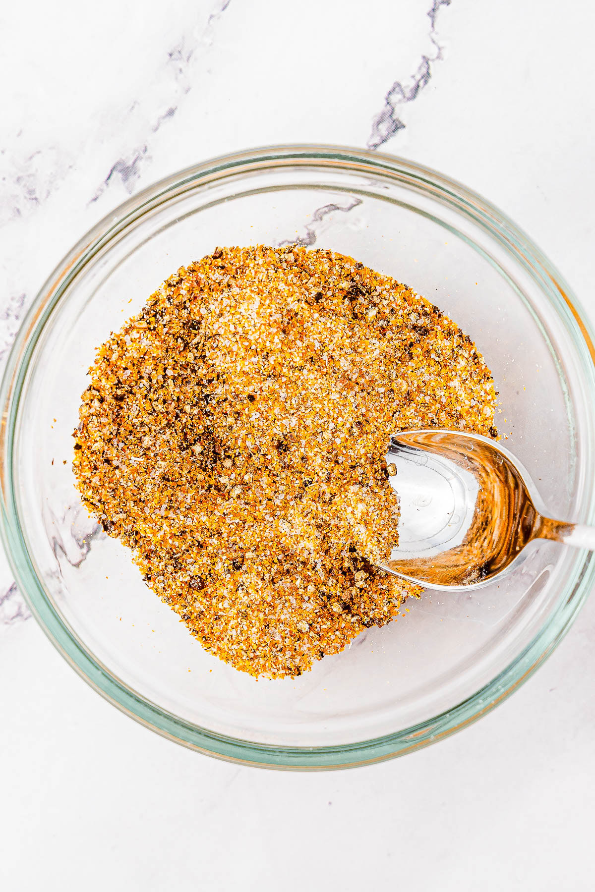 A clear glass bowl filled with a mixed seasoning blend, with a spoon resting inside, on a white marble surface.