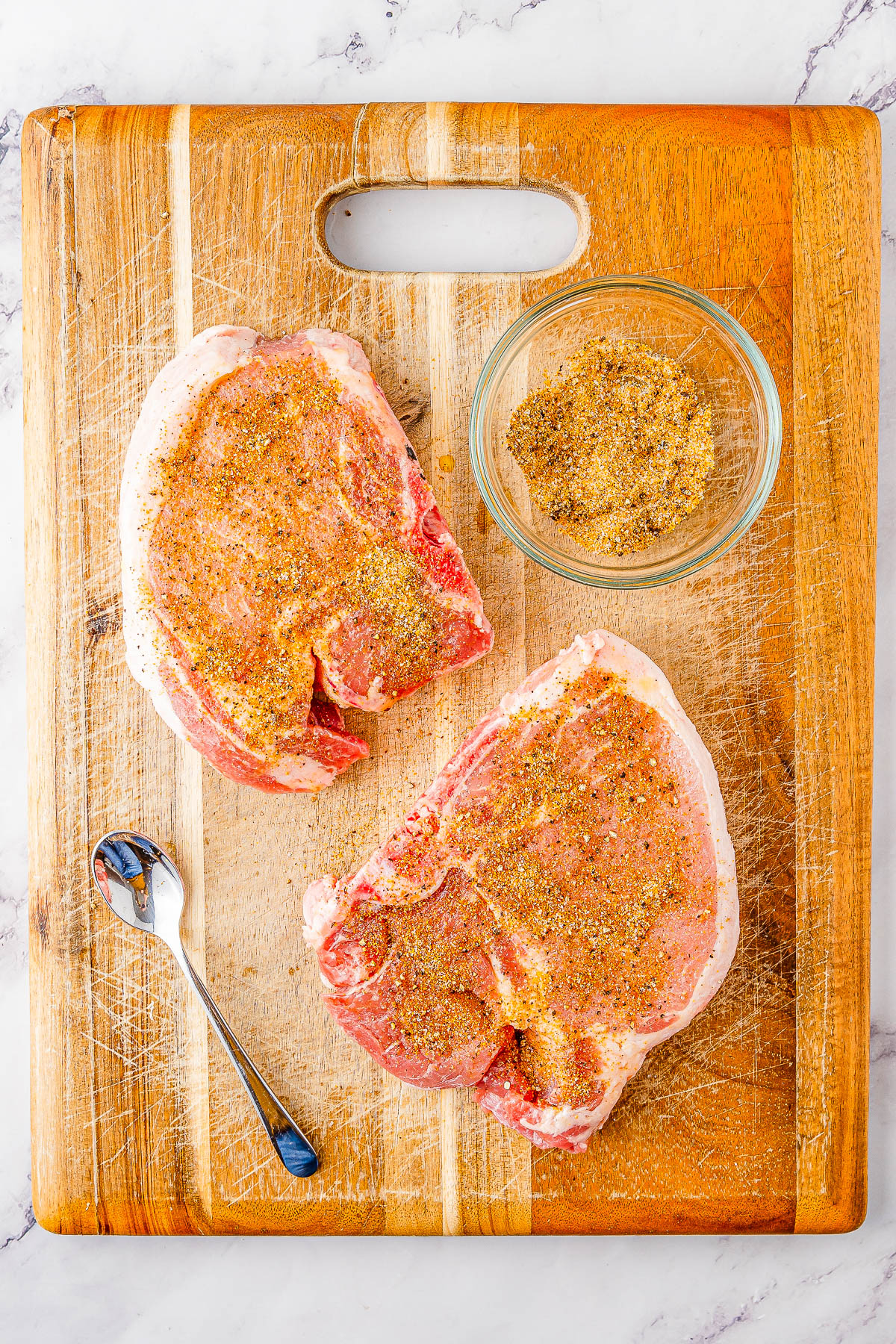 Two raw pork chops on a wooden cutting board are seasoned with a dry rub, with a bowl of seasoning and a spoon placed beside them.