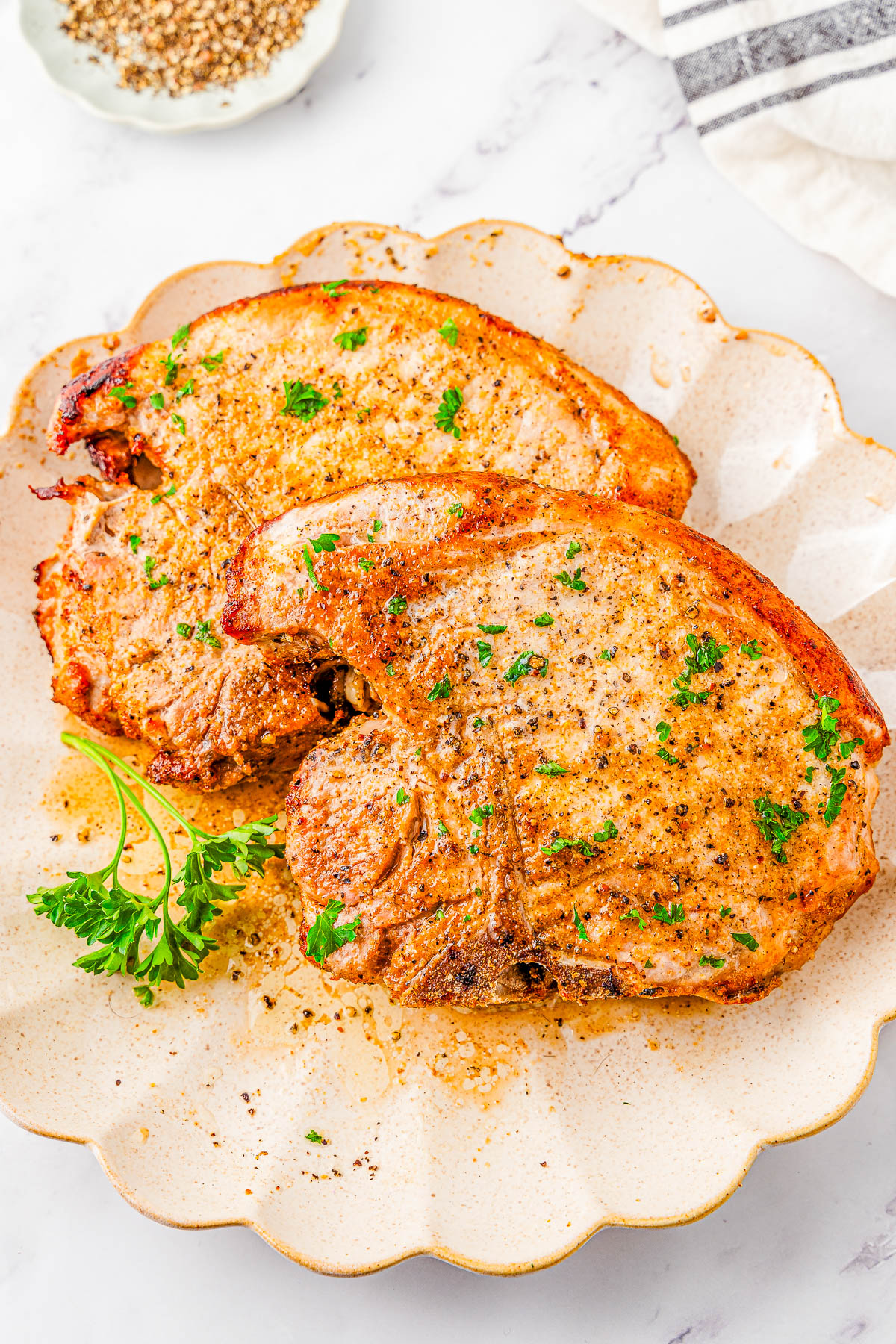 Two cooked, seasoned pork chops garnished with chopped parsley on a beige plate, with a sprig of fresh parsley on the side.