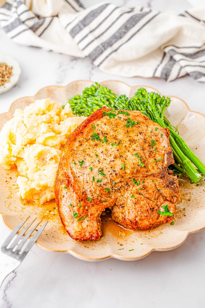 A cooked pork chop served with mashed potatoes and steamed broccolini on a beige plate, with a fork nearby and a striped napkin in the background.