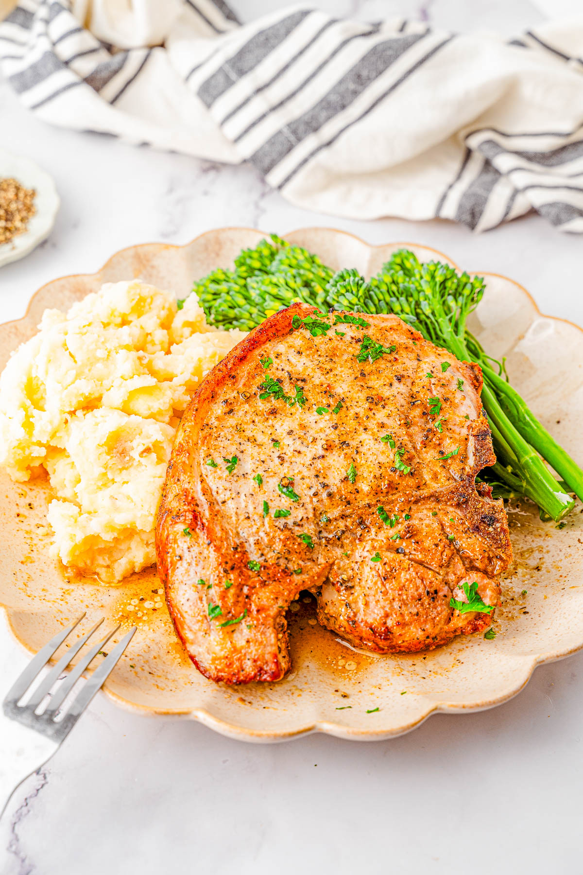 A cooked pork chop served with mashed potatoes and steamed broccolini on a beige plate, with a fork nearby and a striped napkin in the background.