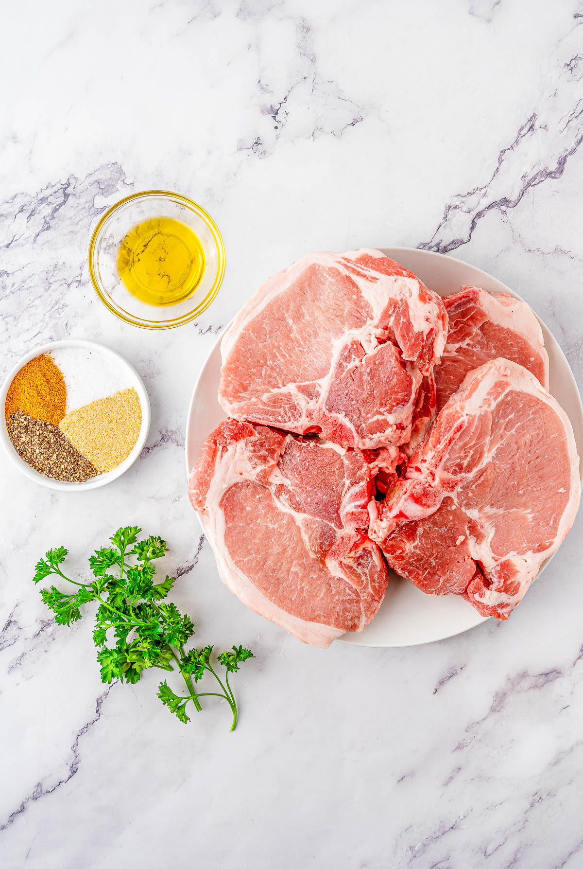 Raw pork chops on a white plate, with a bowl of oil, a small dish of assorted spices, and fresh parsley on a marble surface.
