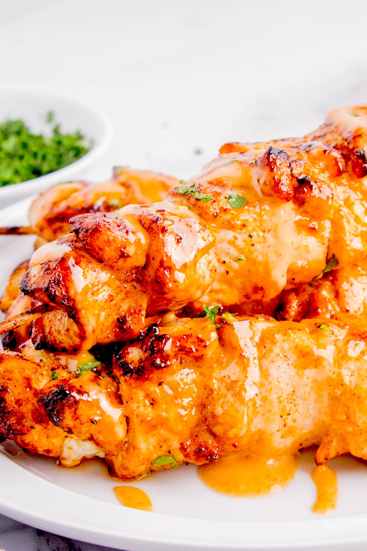 Grilled chicken skewers covered in a creamy orange sauce are served on a white plate, with a small bowl of chopped herbs in the background.