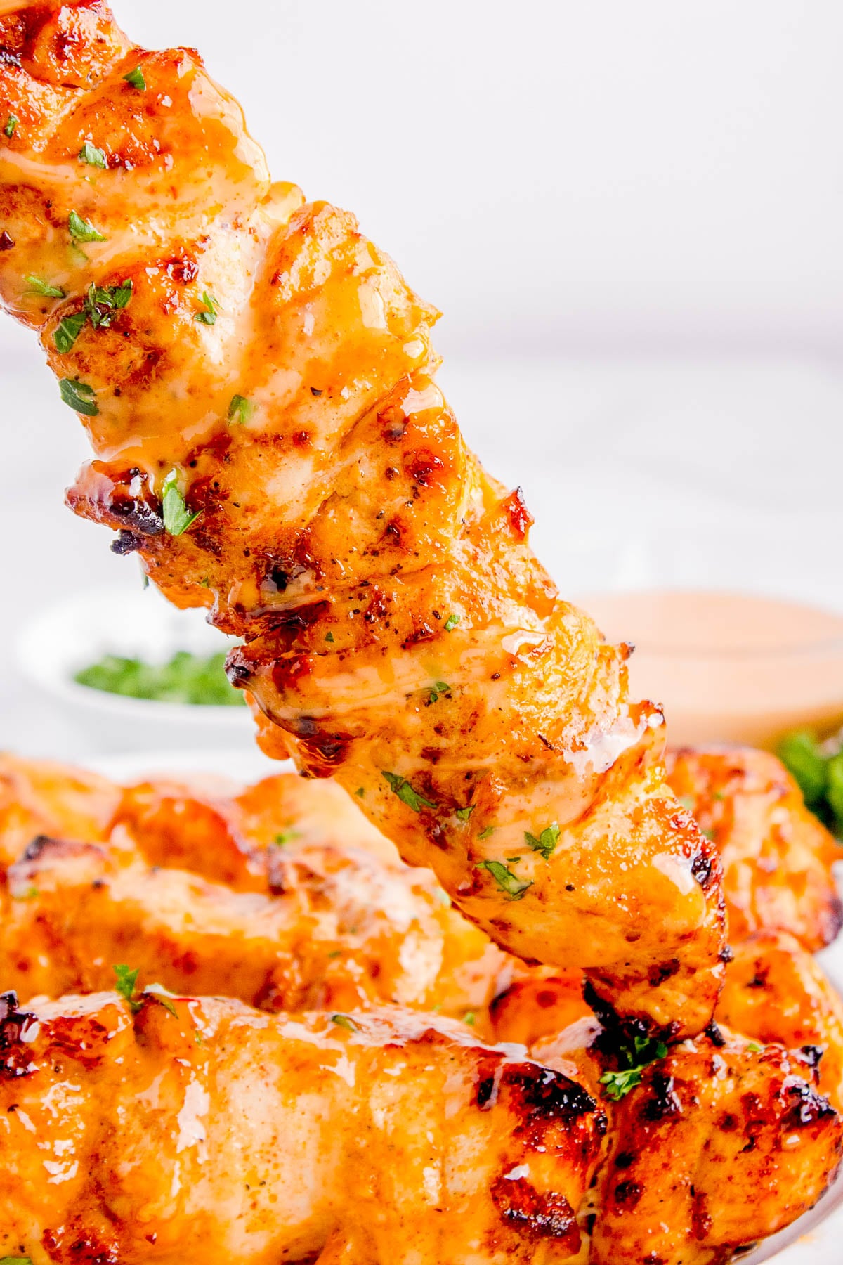 Close-up of a grilled chicken skewer with a golden-brown, slightly charred surface, garnished with herbs, and a dipping sauce in the background.