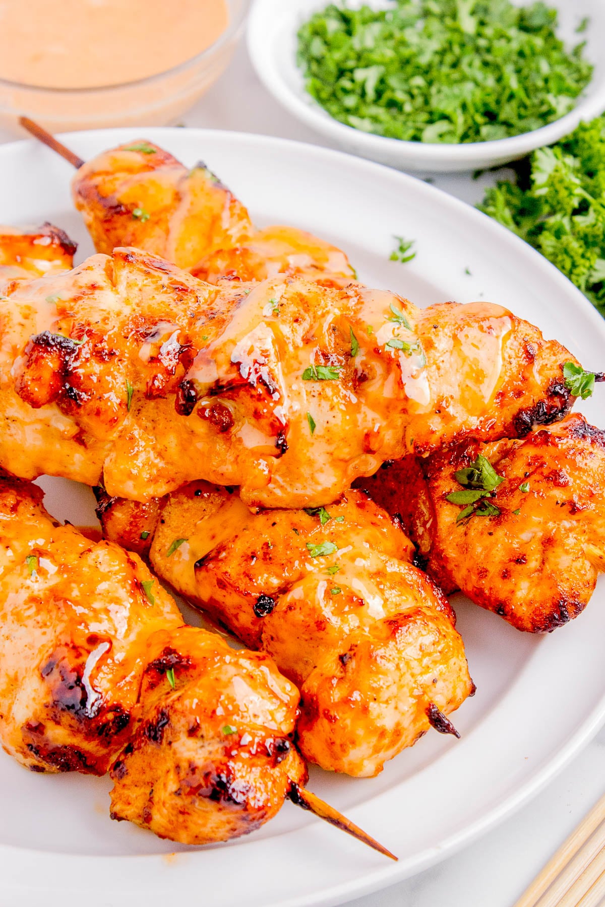 Grilled chicken skewers covered in a glossy orange sauce, garnished with chopped herbs, served on a white plate with fresh parsley in the background.