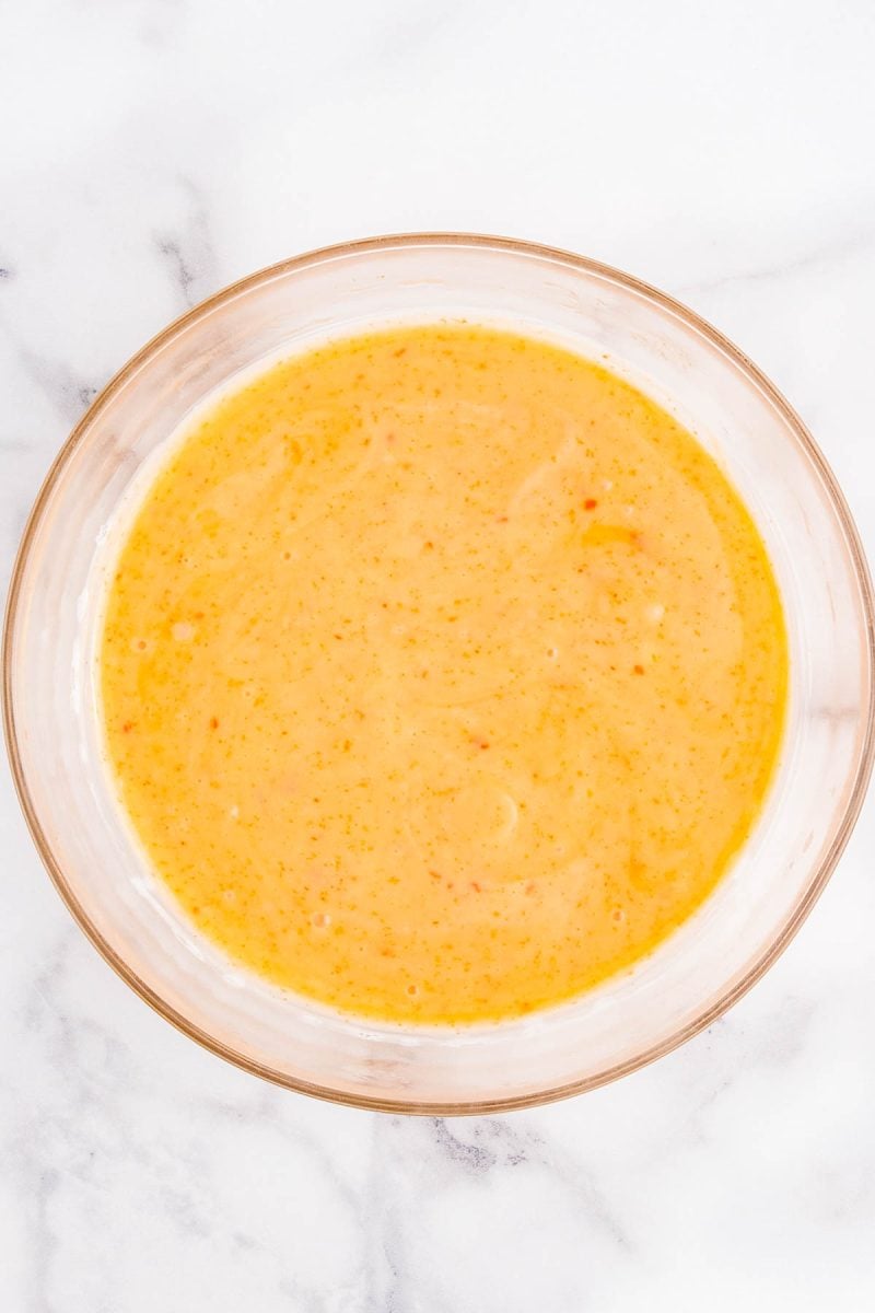 A clear glass bowl filled with a creamy, orange-yellow sauce or dressing sits on a white marble surface.