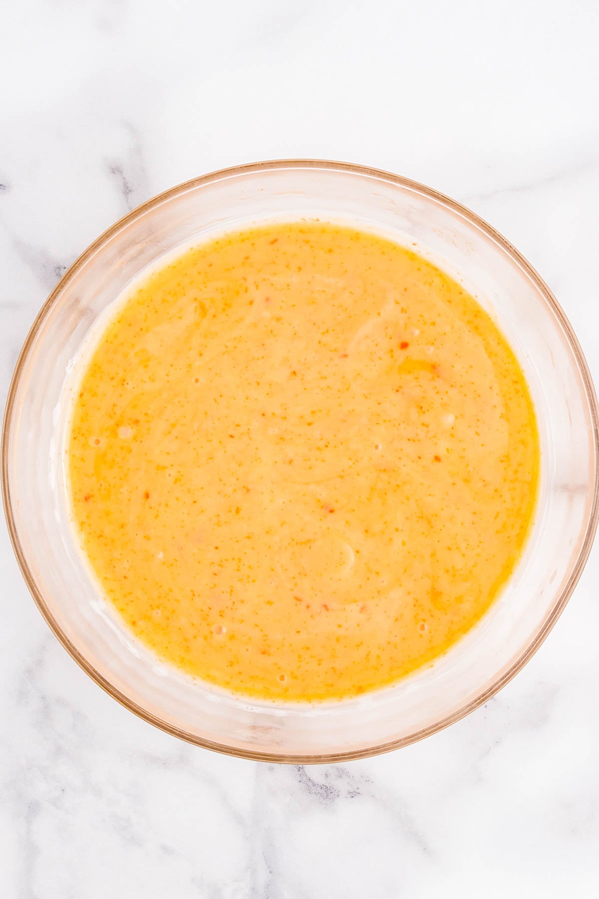 A clear glass bowl filled with a creamy, orange-yellow sauce or dressing sits on a white marble surface.