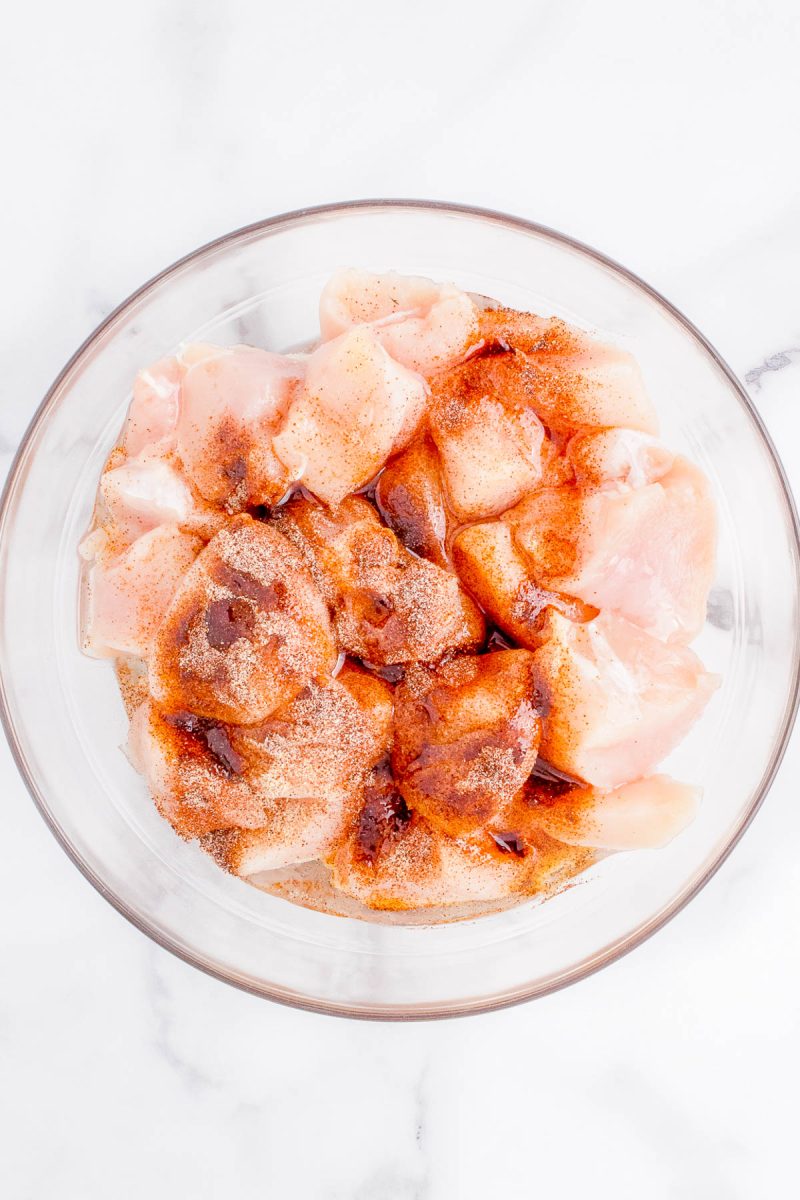 A glass bowl containing raw diced chicken pieces sprinkled with various seasonings on a white marble surface.
