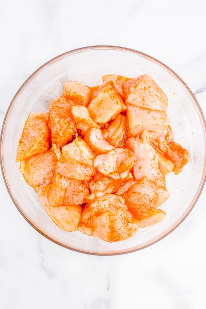 Raw chicken pieces coated in a reddish marinade are arranged in a clear glass bowl on a white marble surface.