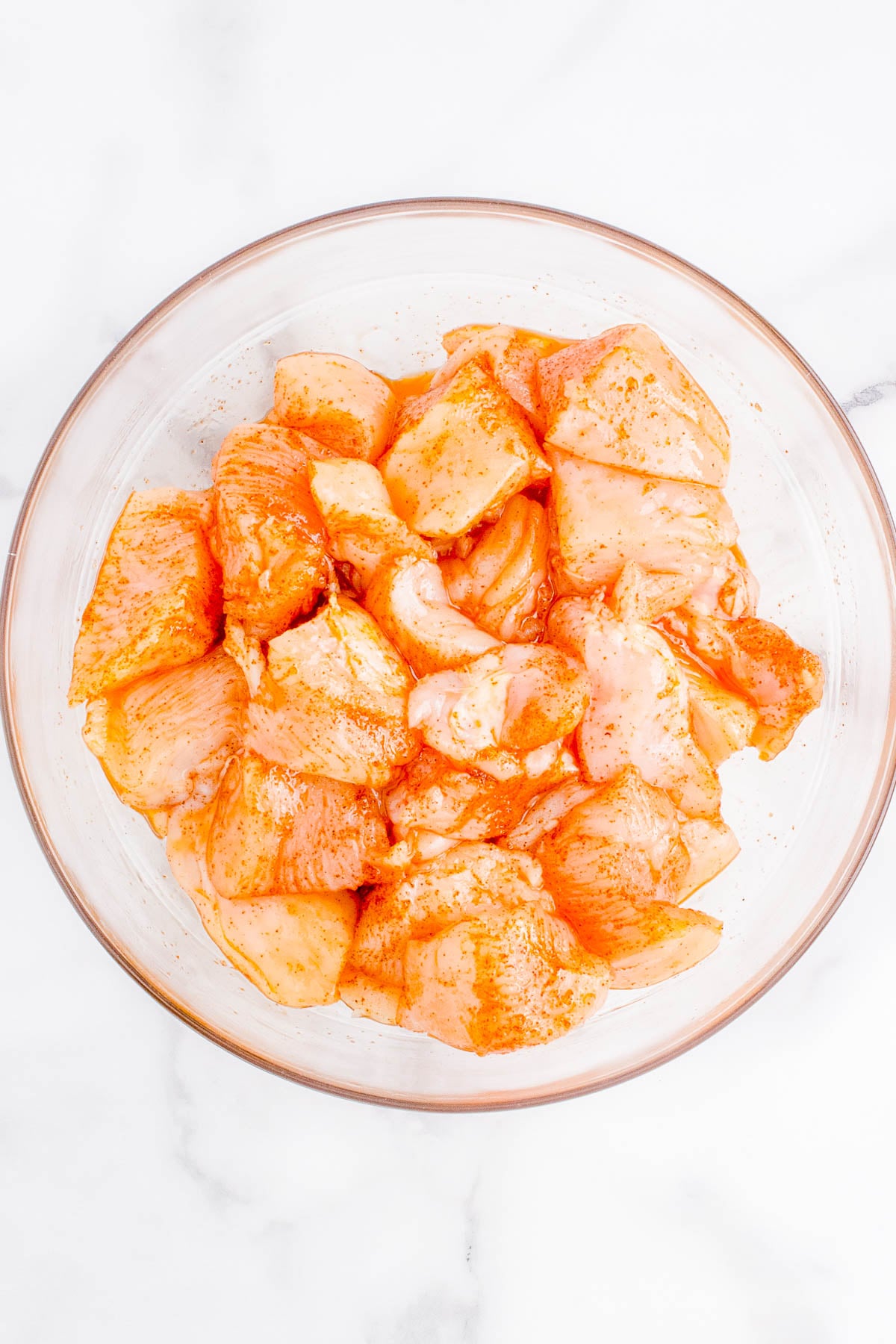 Raw chicken pieces coated in a reddish marinade are arranged in a clear glass bowl on a white marble surface.