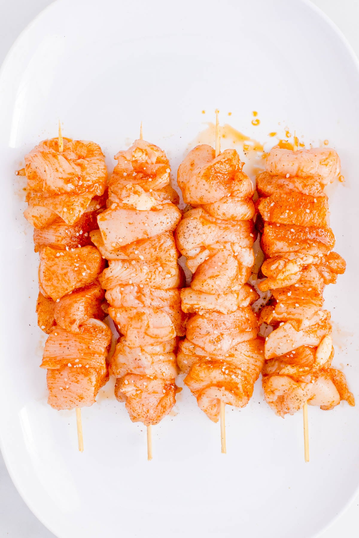 Three skewers of raw, marinated chicken pieces arranged in a row on a white oval plate.