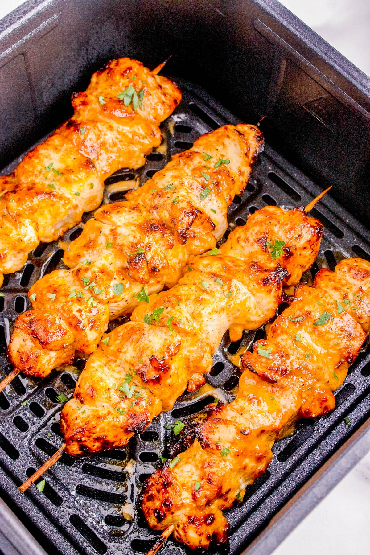Four grilled chicken skewers with golden brown char and sprinkled herbs in an air fryer basket.