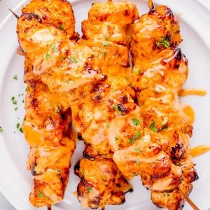 Three grilled chicken skewers drizzled with orange sauce and garnished with chopped herbs, served on a white plate.